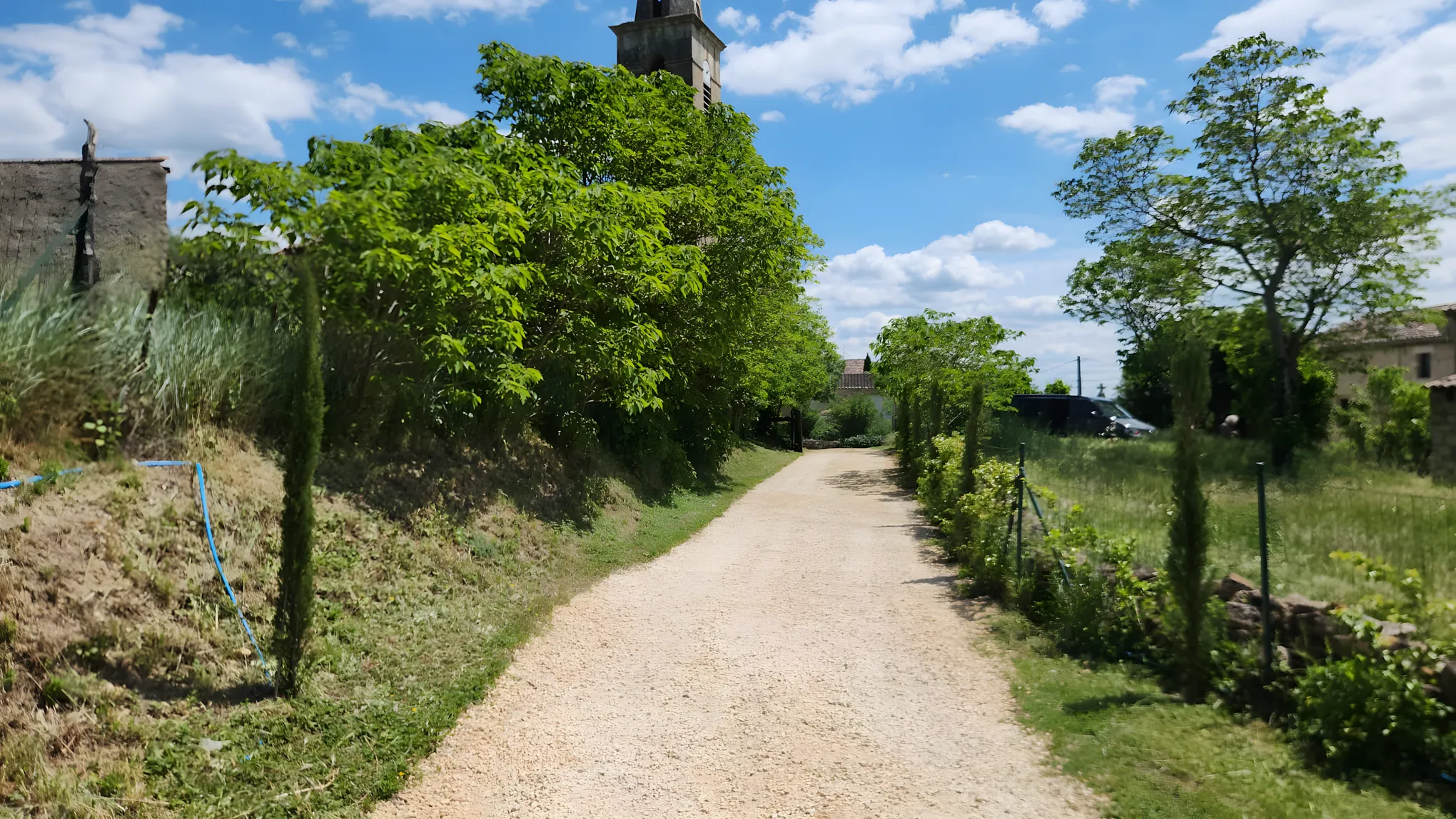 Allée paysagère en gravier bordée de cyprès et d’arbres, clocher du village en arrière-plan