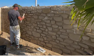 Andy au travail : il sculpte les pierres d’un mur extérieur dans l’enduit à la chaux, chantier en cours